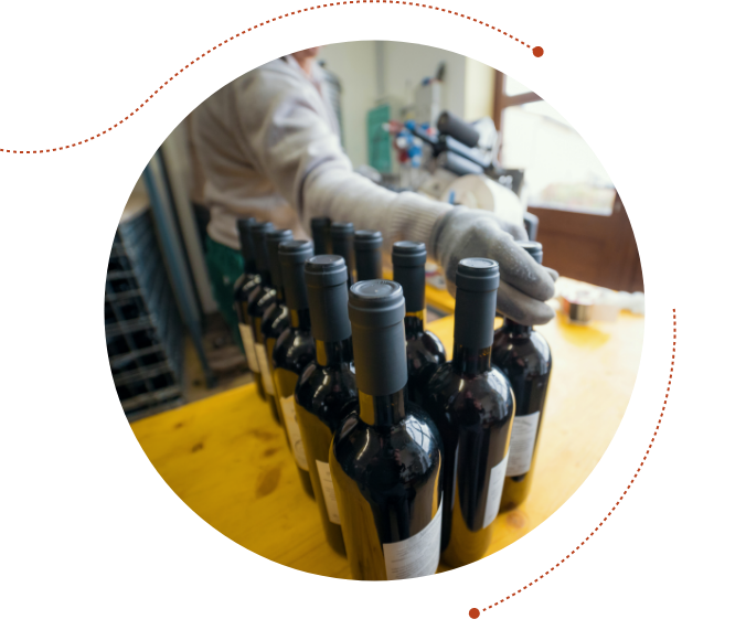 Worker wearing gloves is organizing rows of wine bottles on a yellow table. The bottles are neatly arranged in clusters, and the worker is readying them for packaging.